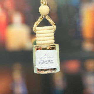 A small glass bottle diffuser or perfume bottle hanging from a rope loop with a wooden bead and cylindrical wooden cap. The bottle has a white label reading 'Wooden Aroma' and 'MY FAVORITE CRYSTAL SHOP', with a warm amber-toned liquid inside.