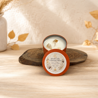 A round tin candle with its lid placed in front, set on a wooden slice tray on a light wood surface. The candle has white wax and a decorative label, with dried botanical elements in the background.