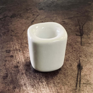 A small glossy white ceramic cup or candle holder with a rounded square shape, placed on a dark brown textured wooden surface.