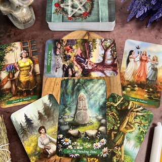 A spread of tarot cards from the Green Witch Tarot deck, featuring colorful illustrations of people, plants, and animals in a natural setting. The cards are placed on a wooden surface with a purple flower in the background.
